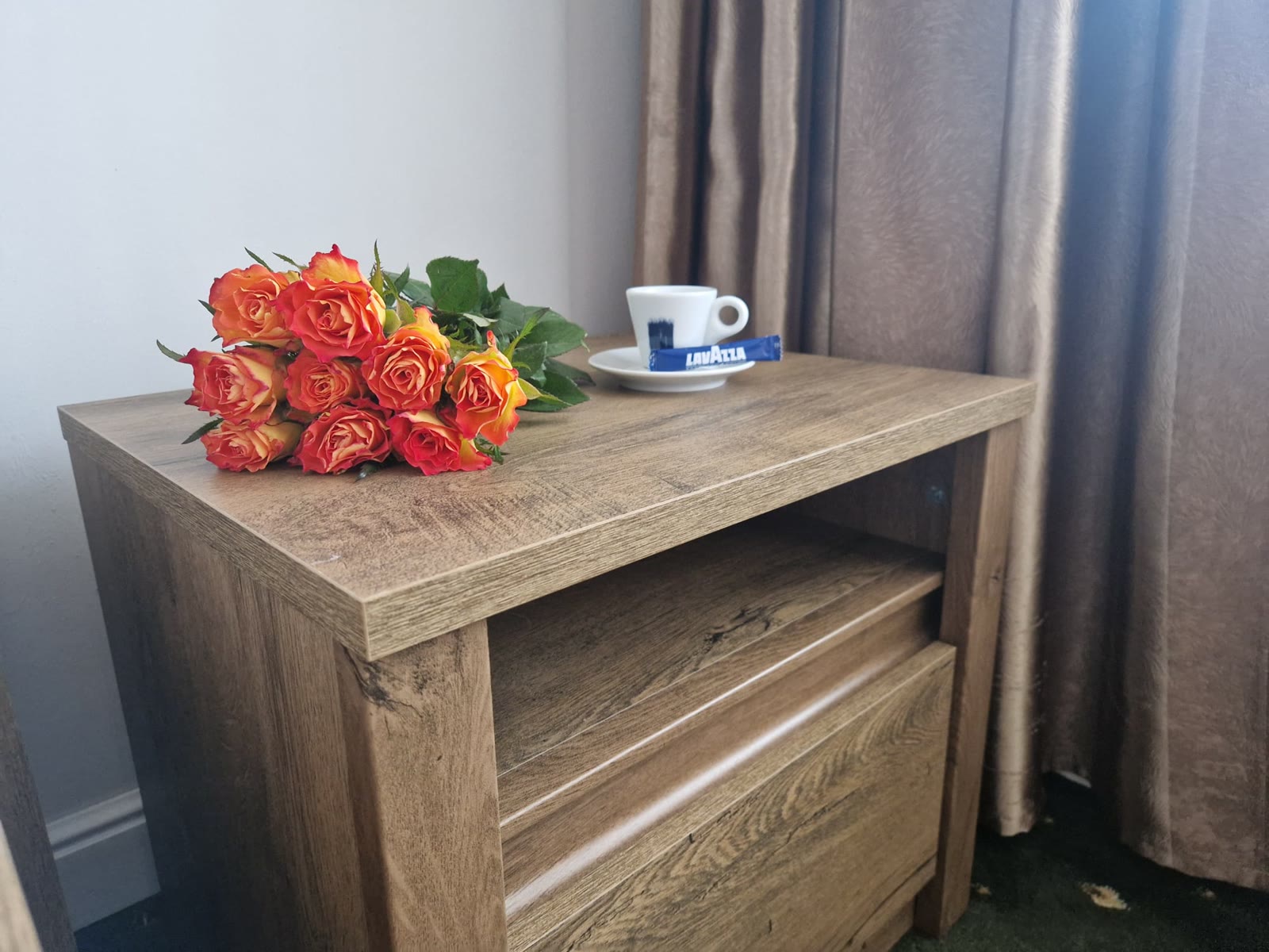 Noptieră din lemn cu un buchet de trandafiri portocalii și o ceașcă de cafea, lângă draperiile camerei