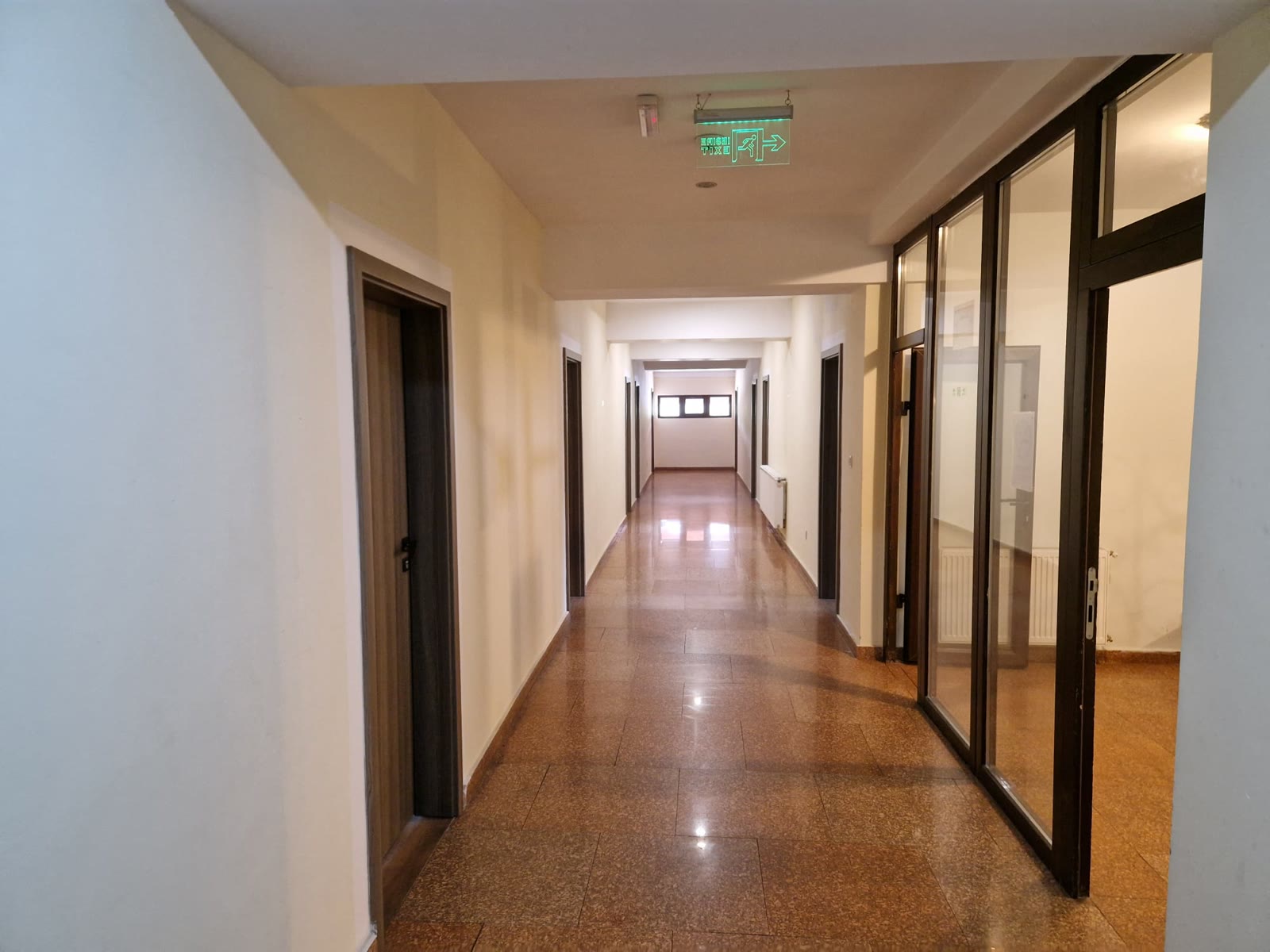 Holul camerelor: pardoseală lucioasă din piatră, uși de lemn de o parte și de alta și un perete despărțitor din sticlă cu rame maro