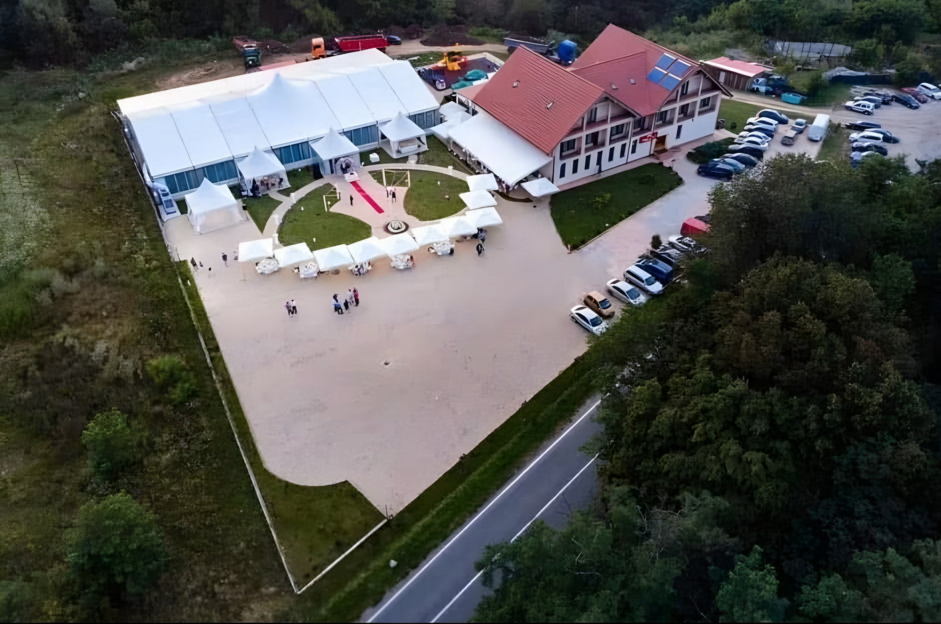 Vedere din dronă: pensiunea Casa cu Tei cu acoperiș roșu, cortul alb de evenimente alături, curtea cu covor roșu și umbrele albe, parcarea și drumul DN58