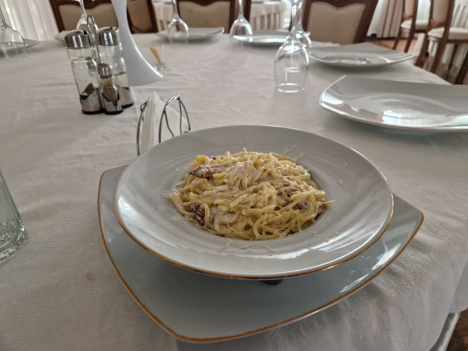 Farfurie cu paste carbonara cu parmezan, servită pe o masă cu față de masă albă, pahare și solnițe, în sala de mese