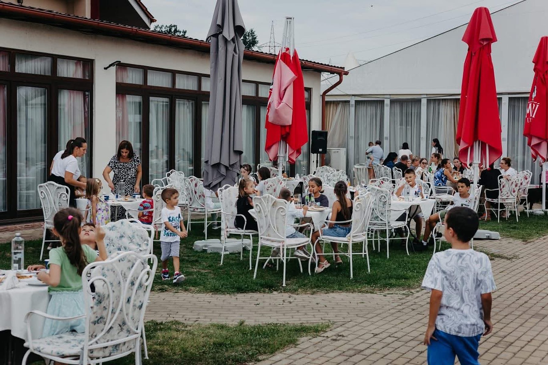 Copii la masă în curtea restaurantului, pe scaune albe din fier forjat, lângă umbrelele roșii