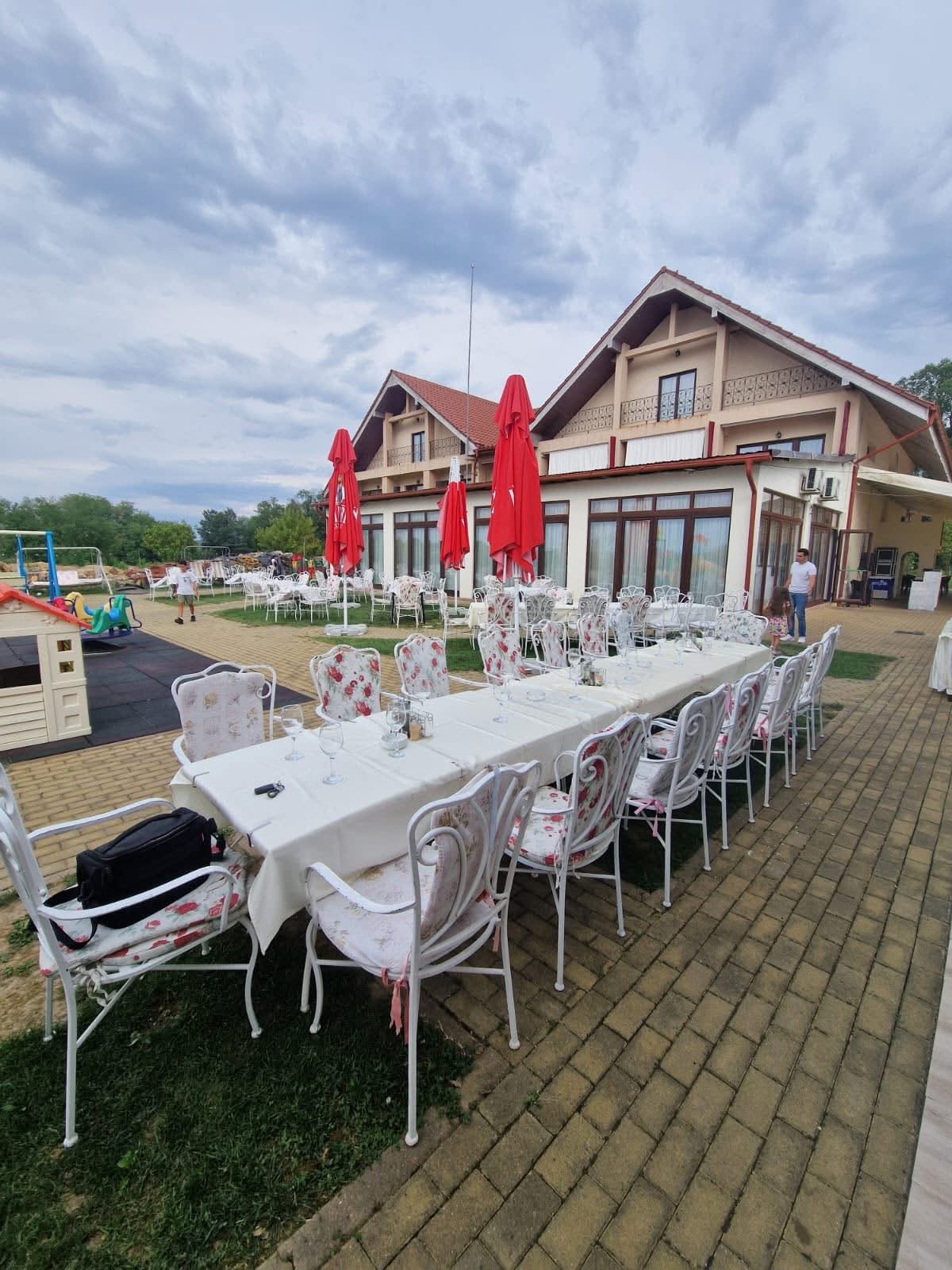 Masă lungă cu fețe de masă albe și scaune din fier forjat cu perne înflorate, în fața pensiunii; în stânga, căsuța și leagănele parcului de joacă